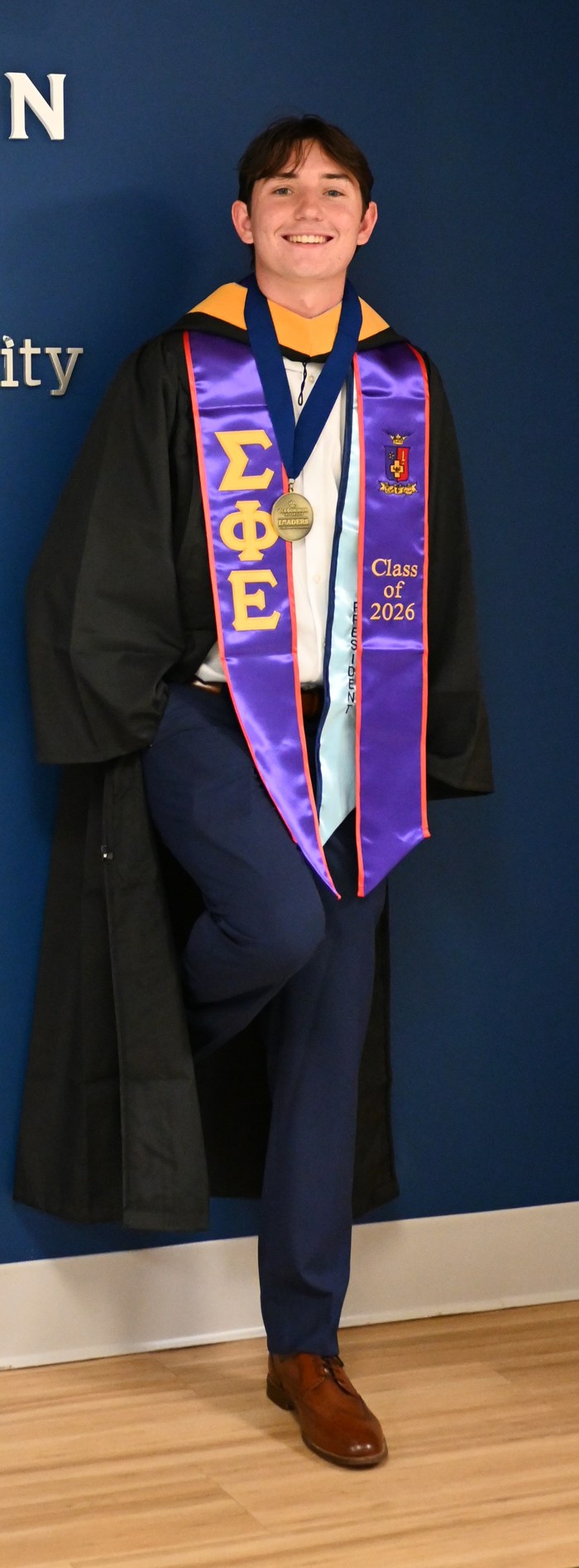 Michael Scholz, in a graduation gown, standing in front of the Old Dominion University School of Cybersecurity sign.